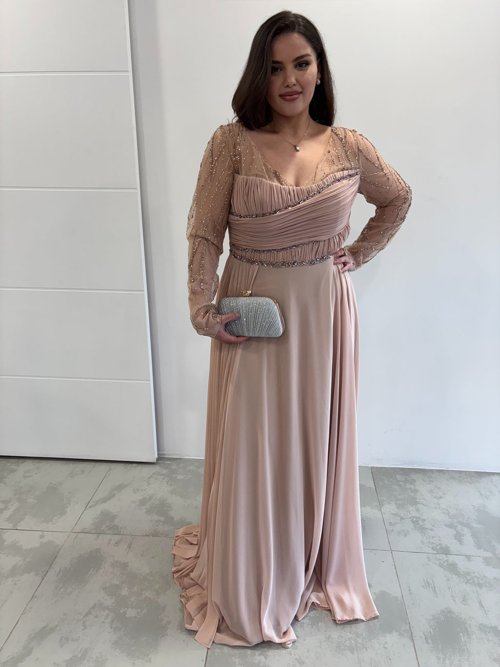 Robe de soirée rose poudrée évasée  vue entière de face, manches longues transparentes pailletées, buste et traine sur le coté plissés 
