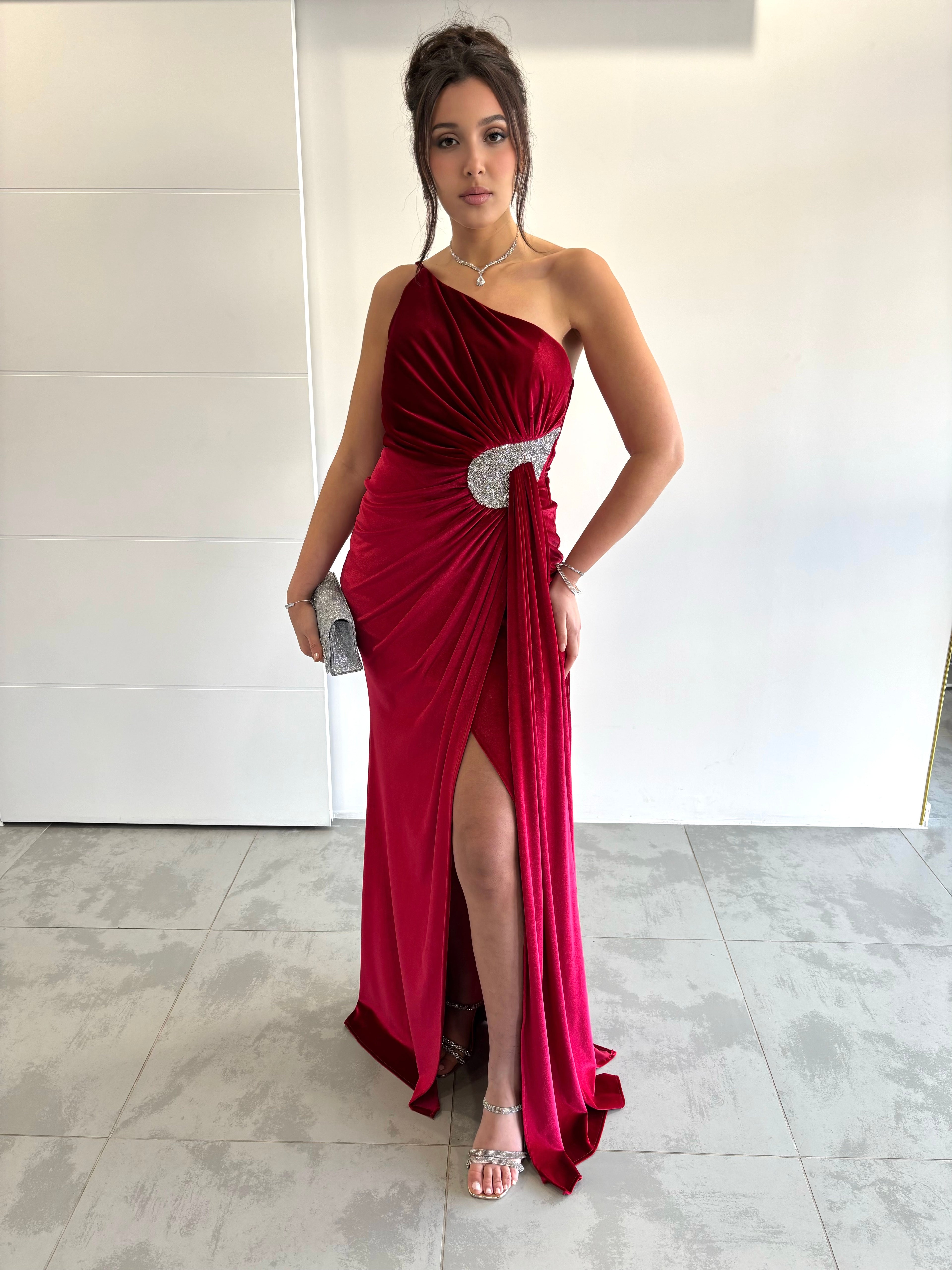 Robe de soirée rouge en velours pailleté avec col asymétrique, détail argenté, fente et traîne sur le côté
